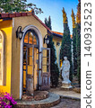 Rustic arched doorway with weathered wooden doors, classical statue and cypress trees on a Mediterranean-style building 140932523