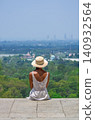 Woman in white dress and straw hat sitting on hilltop viewpoint overlooking tropical city landscape in Thailand 140932564