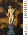 Classical female garden statue holding wheat sheaves on a decorative pedestal against a stone wall in warm evening light 140932569