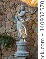 Classical female garden statue holding grapes on a floral pedestal against a rustic stone wall in warm evening light 140932570