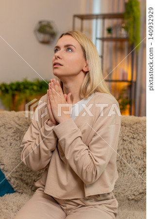 Adult woman praying at home on sofa, folding hands looking upward with sincere emotional expression Adult woman praying at home on sofa, folding hands looking upward with sincere emotional expression 140932929