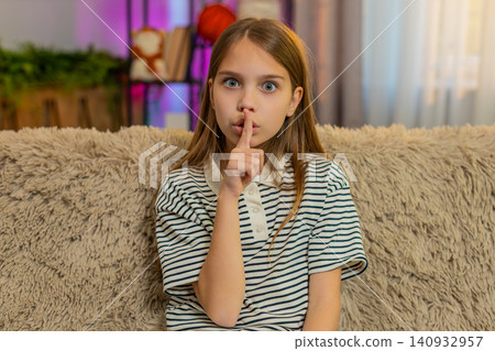 Child girl kid sitting on sofa at home showing silence gesture with finger near lips looking serious 140932957