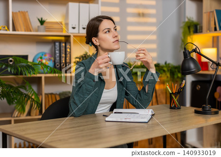 Young woman on home table writing notes removing glasses drinking coffee during study break indoors Young woman on home table writing notes removing glasses drinking coffee during study break indoors 140933019