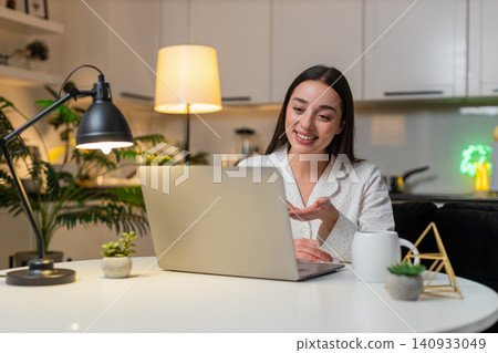 Young woman on laptop webcam waves hello speaks shows charts to client listens nods online evening 140933049