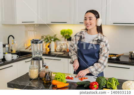 Young woman cutting cherry tomatoes listening to music preparing fresh vegetarian salad at home meal Young woman cutting cherry tomatoes listening to music preparing fresh vegetarian salad at home meal 140933110