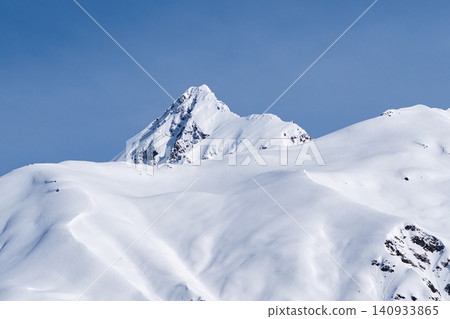 A mountain range covered with a deep layer of white snow is captured against a cloudless bright blue sky. A mountain range covered with a deep layer of white snow is captured against a cloudless bright blue sky. 140933865