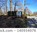 The entrance to the Lakewinds residential complex features a white sign with silhouettes of ducks. Tall trees surround the entrance, and an empty road lies to the right 140934076