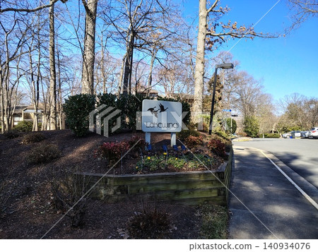 The entrance to the Lakewinds residential complex features a white sign with silhouettes of ducks. Tall trees surround the entrance, and an empty road lies to the right The entrance to the Lakewinds residential complex features a white sign with silhouettes of ducks. Tall trees surround the entrance, and an empty road lies to the right 140934076