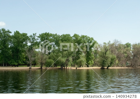 A tranquil landscape featuring lush green foliage by the Volga River, with the surrounding scenery clearly reflected in the water; natural lighting A tranquil landscape featuring lush green foliage by the Volga River, with the surrounding scenery clearly reflected in the water; natural lighting 140934278