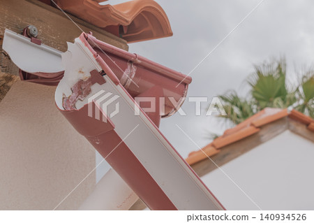 Close up shot of a broken and disconnected plastic rain gutter on a tiled residential house roof edge needing repair work under a cloudy sky 140934526