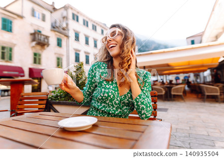 Happy woman sitting by table drinking coffee of outdoor sidewalk cafe restaurant, on summer day. 140935504