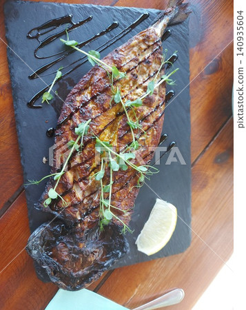 Grilled fish served on a slate plate, in wooden table, with grilled fish, lemon wedge, herbs, dark sauce, with slate plate, wooden table texture, from a top-down view perspective, clear detail. Grilled fish served on a slate plate, in wooden table, with grilled fish, lemon wedge, herbs, dark sauce, with slate plate, wooden table texture, from a top-down view perspective, clear detail. 140935604