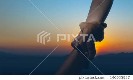 Silhouette of two hands holding each other firmly for support, rescue, and teamwork against a beautiful sunset sky background Silhouette of two hands holding each other firmly for support, rescue, and teamwork against a beautiful sunset sky background 140935937