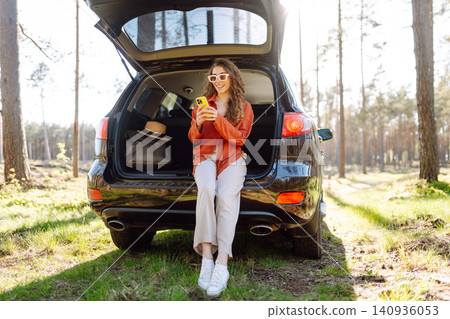 Beautiful woman sits in the trunk of a car outdoor and uses a smartphone. Active life. Beautiful woman sits in the trunk of a car outdoor and uses a smartphone. Active life. 140936053