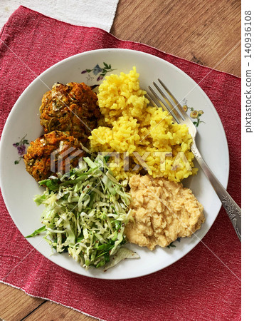 A plate holds turmeric rice, fresh salad, hummus, and two carrot and zucchini meatballs. This meal offers a healthy option for breakfast or lunch enjoyed at the dining table A plate holds turmeric rice, fresh salad, hummus, and two carrot and zucchini meatballs. This meal offers a healthy option for breakfast or lunch enjoyed at the dining table 140936108