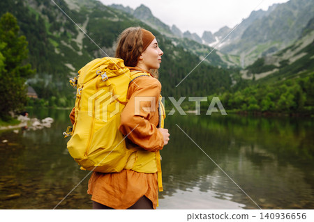 Female traveler with hiking backpack poses against backdrop of mountain lake. Adventure concept. Female traveler with hiking backpack poses against backdrop of mountain lake. Adventure concept. 140936656