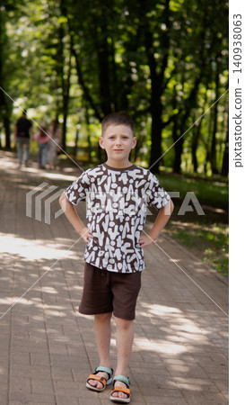 Full length portrait of young boy standing with hands on hips on sun dappled paved park path surrounded by tall leafy trees, wearing patterned short sleeve Full length portrait of young boy standing with hands on hips on sun dappled paved park path surrounded by tall leafy trees, wearing patterned short sleeve 140938063
