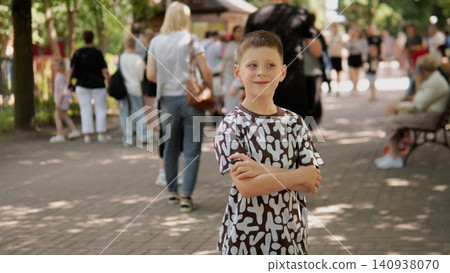Smiling school-age boy stands with folded arms on tree-lined park walkway, wearing patterned short-sleeve shirt and shorts while people stroll and sit on Smiling school-age boy stands with folded arms on tree-lined park walkway, wearing patterned short-sleeve shirt and shorts while people stroll and sit on 140938070