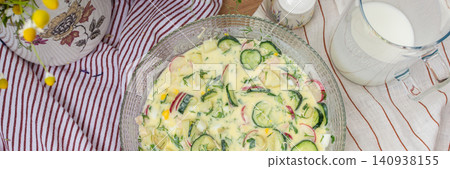 A bowl of okroshka filled with potatoes, chicken breast, cucumbers, herbs, and boiled eggs sits on a table. Nearby, a floral vase adds color to the setting, banner 140938155
