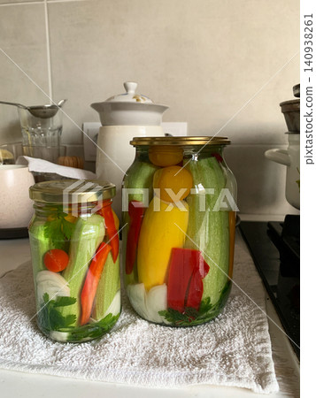Jars filled with cucumbers, peppers, and zucchini sit on a wooden countertop. Fresh vegetables and herbs surround the jars as people prepare food for winter storage in a kitchen Jars filled with cucumbers, peppers, and zucchini sit on a wooden countertop. Fresh vegetables and herbs surround the jars as people prepare food for winter storage in a kitchen 140938261