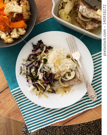 A plate contains rice, baked hake, and fresh vegetables. A salad with various colors is next to it. This setting shows a simple meal on a wooden table, suitable for breakfast or lunch A plate contains rice, baked hake, and fresh vegetables. A salad with various colors is next to it. This setting shows a simple meal on a wooden table, suitable for breakfast or lunch 140938272