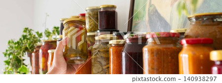 Canned vegetables and fruits are lined up on a shelf in a kitchen, showing winter preparations. A painting of a basket is seen in the background as part of the decor, banner Canned vegetables and fruits are lined up on a shelf in a kitchen, showing winter preparations. A painting of a basket is seen in the background as part of the decor, banner 140938297