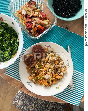 A plate holds green buckwheat mixed with carrots and zucchini alongside beef meatballs. Fresh salad and a bowl of berries sit nearby on a table A plate holds green buckwheat mixed with carrots and zucchini alongside beef meatballs. Fresh salad and a bowl of berries sit nearby on a table 140938309