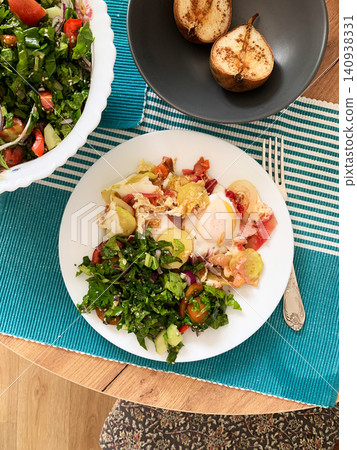 A meal of boiled potatoes, an omelette, stewed vegetables, and fresh salad is placed on a plate on a wooden table. There are sliced pears in a bowl nearby A meal of boiled potatoes, an omelette, stewed vegetables, and fresh salad is placed on a plate on a wooden table. There are sliced pears in a bowl nearby 140938331