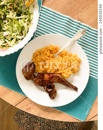 A meal is served on a white plate, showing rice colored with turmeric and pieces of baked rabbit beside a fresh salad in a bowl. The meal is set on a wooden table with a blue cloth A meal is served on a white plate, showing rice colored with turmeric and pieces of baked rabbit beside a fresh salad in a bowl. The meal is set on a wooden table with a blue cloth 140938349