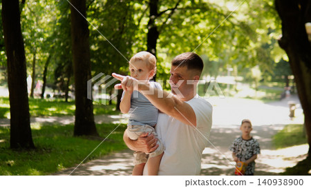 Young father holding toddler in arms while pointing toward park path lined with sunlit trees, shaded lawn, and dappled sunlight; child looks toward indicated 140938900