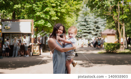 Young mother cradling toddler son while standing on sunlit park path lined with green trees and evergreen shrubs; relaxed family scene with casual summer Young mother cradling toddler son while standing on sunlit park path lined with green trees and evergreen shrubs; relaxed family scene with casual summer 140938903