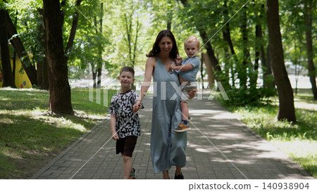 Smiling mother in long blue dress walking down tree-lined paved park path while holding toddler on hip and older child by hand, dappled sunlight through green Smiling mother in long blue dress walking down tree-lined paved park path while holding toddler on hip and older child by hand, dappled sunlight through green 140938904