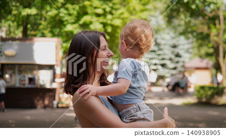 Young mother lifts toddler boy in arms while both smile, outdoor park scene with sunlit trees and blurred background buildings, casual summer clothing, warm Young mother lifts toddler boy in arms while both smile, outdoor park scene with sunlit trees and blurred background buildings, casual summer clothing, warm 140938905