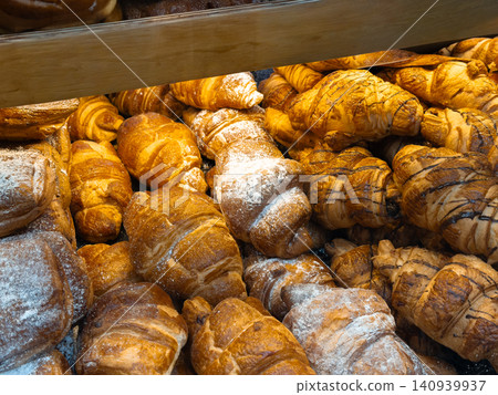 Fresh golden croissants stacked inside a bakery display case, featuring a variety of buttery flaky pastries dusted with sugar and drizzled with chocolate for gourmet treats Fresh golden croissants stacked inside a bakery display case, featuring a variety of buttery flaky pastries dusted with sugar and drizzled with chocolate for gourmet treats 140939937