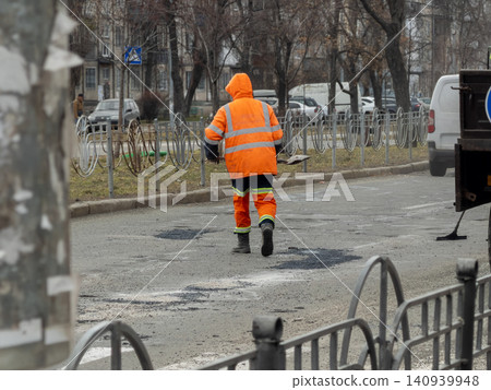 A construction worker in high visibility gear repairs road potholes with a shovel on a busy city street to ensure essential infrastructure maintenance during the bright daylight A construction worker in high visibility gear repairs road potholes with a shovel on a busy city street to ensure essential infrastructure maintenance during the bright daylight 140939948