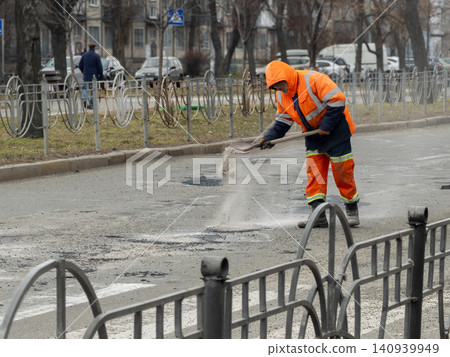 A dedicated road worker in a bright orange uniform uses a shovel to fill a deep pothole with gravel on an urban street to support city infrastructure maintenance and traffic safety A dedicated road worker in a bright orange uniform uses a shovel to fill a deep pothole with gravel on an urban street to support city infrastructure maintenance and traffic safety 140939949
