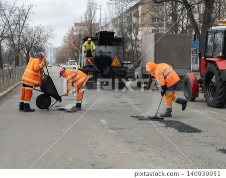 Professional road crew workers in bright orange uniforms repair asphalt on a busy city street using heavy machinery and manual tools for essential urban infrastructure maintenance Professional road crew workers in bright orange uniforms repair asphalt on a busy city street using heavy machinery and manual tools for essential urban infrastructure maintenance 140939951