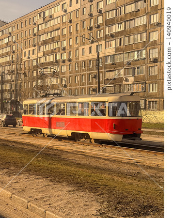 Retro red tram moving along tracks past a multi-story apartment block in a city, representing public transportation efficiency and the nostalgic charm of daily urban commuting life Retro red tram moving along tracks past a multi-story apartment block in a city, representing public transportation efficiency and the nostalgic charm of daily urban commuting life 140940019