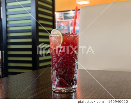 Cold red berry cocktail with ice cubes and a fresh lime garnish in a faceted highball glass, sitting on a dark wooden table in a cozy cafe setting to evoke refreshment and leisure Cold red berry cocktail with ice cubes and a fresh lime garnish in a faceted highball glass, sitting on a dark wooden table in a cozy cafe setting to evoke refreshment and leisure 140940020