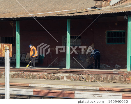 Two cleaners in reflective safety vests sweep the dusty platform of an old railway station, highlighting the daily maintenance work performed in public transportation infrastructure Two cleaners in reflective safety vests sweep the dusty platform of an old railway station, highlighting the daily maintenance work performed in public transportation infrastructure 140940030