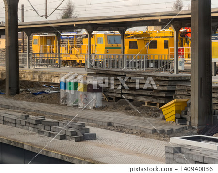 A bright yellow railway maintenance train parked at a construction station platform with colorful recycling bins and paving stones, illustrating industrial urban infrastructure A bright yellow railway maintenance train parked at a construction station platform with colorful recycling bins and paving stones, illustrating industrial urban infrastructure 140940036