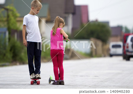 Cute young blond children girl in pink clothing on scooter and handsome boy on skateboard playing on sunny street on blurred summer bright background. Children activities, games and fun concept. Cute young blond children girl in pink clothing on scooter and handsome boy on skateboard playing on sunny street on blurred summer bright background. Children activities, games and fun concept. 140940280