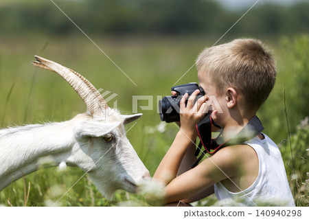 Profile portrait of young blond cute handsome child boy taking picture of funny curious goat looking straight in camera on bright sunny summer day on blurred light green grassy copyspace background. Profile portrait of young blond cute handsome child boy taking picture of funny curious goat looking straight in camera on bright sunny summer day on blurred light green grassy copyspace background. 140940298