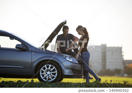 Young couple, handsome man and attractive woman at car with popped hood checking oil level in engine using dipstick on clear sky background. Transportation, vehicles problems and breakdowns concept. Young couple, handsome man and attractive woman at car with popped hood checking oil level in engine using dipstick on clear sky background. Transportation, vehicles problems and breakdowns concept. 140940302