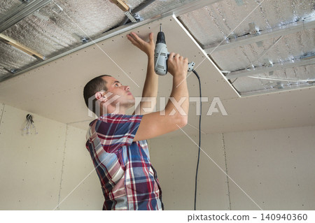 Young man in usual clothing and work gloves fixing drywall suspended ceiling to metal frame using electrical screwdriver on ceiling insulated with shiny aluminum foil. DIY, do it yourself concept. Young man in usual clothing and work gloves fixing drywall suspended ceiling to metal frame using electrical screwdriver on ceiling insulated with shiny aluminum foil. DIY, do it yourself concept. 140940360