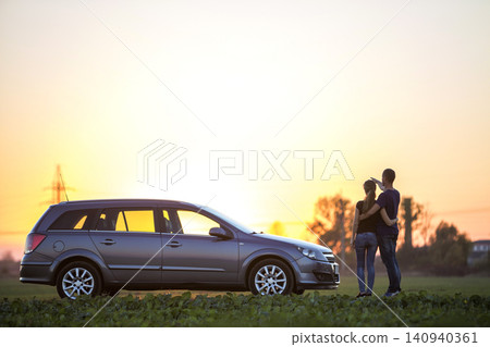 Young couple, slim attractive woman with long ponytail and handsome man standing at silver car in green field on clear sky at sunset or sunrise copy space background. Young couple, slim attractive woman with long ponytail and handsome man standing at silver car in green field on clear sky at sunset or sunrise copy space background. 140940361