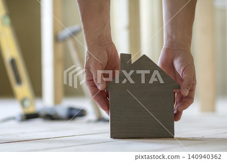 Close-up of muscular male hands holding small brown wooden model house on blurred background of building tools. Construction, investments in real estate property and ownership of dream home concept. Close-up of muscular male hands holding small brown wooden model house on blurred background of building tools. Construction, investments in real estate property and ownership of dream home concept. 140940362