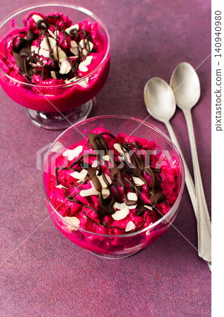 Two bowls of healthy ice cream sit on a table. The ice cream features strawberries, bananas, beets, and Greek yogurt. Chocolate and nuts top the creamy dessert. Spoons are beside the bowls Two bowls of healthy ice cream sit on a table. The ice cream features strawberries, bananas, beets, and Greek yogurt. Chocolate and nuts top the creamy dessert. Spoons are beside the bowls 140940980