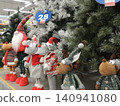 Soft Christmas toys in New Year's clothes and red caps are displayed in a store against a backdrop of fluffy artificial Christmas trees. 140941080