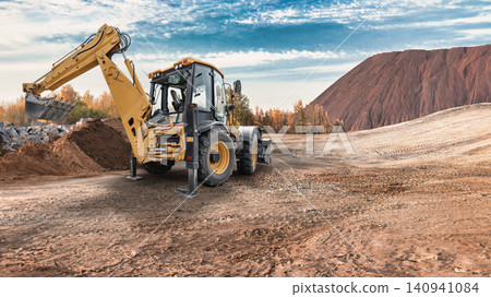 A universal excavator loader is digging and moving dirt at a construction site. The machine is surrounded by a landscape with trees and hills in the background A universal excavator loader is digging and moving dirt at a construction site. The machine is surrounded by a landscape with trees and hills in the background 140941084